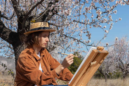 Young men artist painting on canvas in a flower field wearing a hat on a spring dayの写真素材