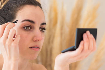 Portrait of a woman plucking her eyebrows with a tweezer looking at mirrorの写真素材