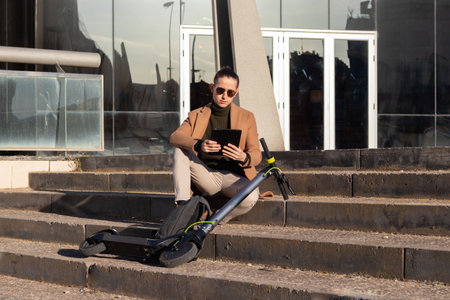 Young handsome bussinesman sitting on stairs in financial district with electric scooter that uses to commute to workの写真素材