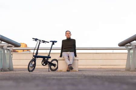 Young man sitting on a park bench with his battery bike on the side looking at the camera smiling with copy spaceの写真素材