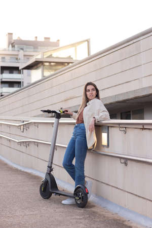 Vertical view of young woman leaning resting with her electric scooter wating for her friend to come with sun behindの写真素材