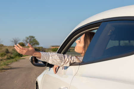 Woman with eyes closed leaning out of car window with arm outside enjoying the fresh breeze of nature in a road tripの写真素材