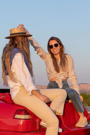 Vertical phoot of two friends enjoying spring break roadtrip sitting on a red car looking at sunset under blue skiesの写真素材