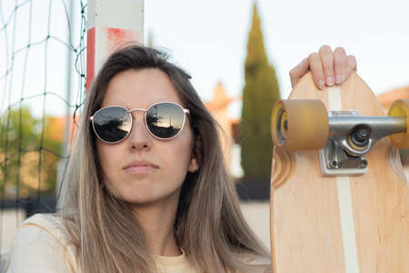 Portrait of a beautiful Hispanic girl seated with a pink skate and the reflection of the sunset on her sunglassesの写真素材