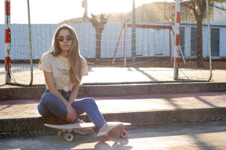 Young latin woman sitting on a stair step relaxed at the park with a skate board under her leg on...