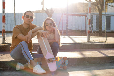 Two hipster friends sitting on steps leaning on cute skate board in a spring afeternoon with sunlight behind themの写真素材