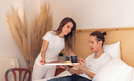 Cute young heterosexual couple on bed about to have yummy breakfast together on a boho minimal bedroomの写真素材