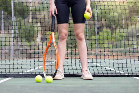 Front view of unrecognizable tennis player woman legs and hands in front of a net holding a ball and a racket on a courtの写真素材