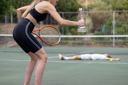 Side view of unrecognizable woman winner of championship holding cup on hand and man looser lying on...