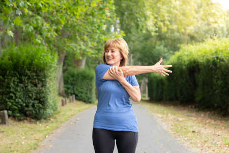 Mature woman in workout outfit smiling to the side streching her arm in the green park and trees aroundの写真素材
