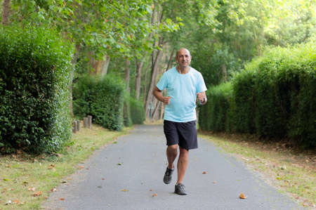 Front portrait in landscape format of a senior man running along a path among the green trees of a...