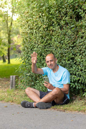 Friendly mature man sitting in the field and waving to the camera while listening to music on his phoneの写真素材