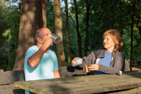 Man and woman sitting on a bench rehydrating with water in a metal cup after trekking in the mountainsの写真素材