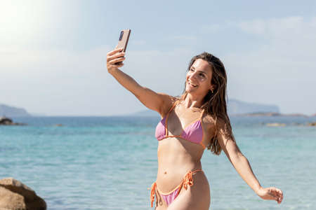 Gorgeous young brunette girl taking a selfie with the beach in the background on a colorful bikini swimwearの写真素材