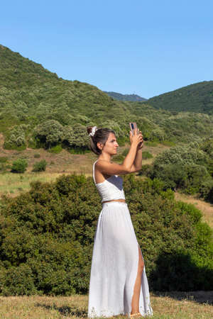 Vertical view of young woman tourist in white outfit top and long skirt taking a picture with her smartphone in summerの写真素材