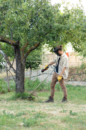 Back of farmer young with unrecognizable faceman working hard and mowing the grass with a brushcutterの写真素材