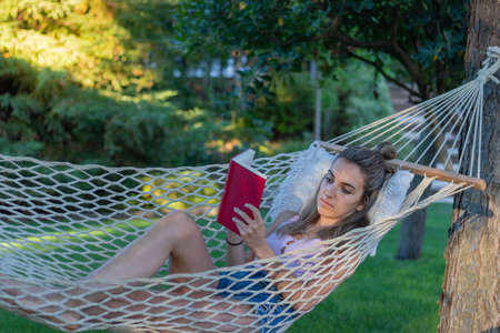 Full lenght photo of young woman relaxing reading a book as leisure activity in a hammock in springtime in the afternoonの写真素材
