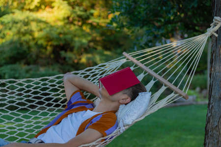 Young man resting and sleeping in a hammock in spring time with a red book covering his eyes and faceの写真素材