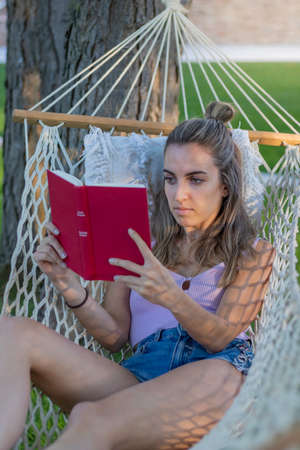 Vertical view of young beautiful woman sitting in hammock and reading a book in a camping enviromentの写真素材