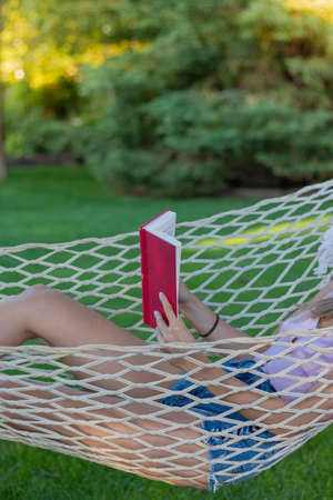 Vertical view of unrecognizable young woman in summer outfit reading a red book with copy space in the gardenの写真素材