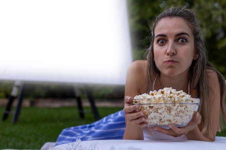 Portrait of a young woman with her eyes open and a face of surprise as her mouth is full of popcorn at the summer cinemaの写真素材