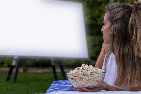 Side view of unrecognizable blonde woman watching a movie in the garden with a projector and popcornsの写真素材