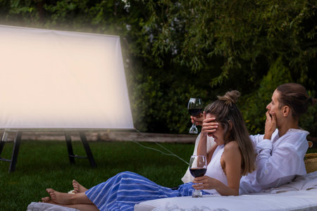 Young couple watching a horror movie in the city park at night surprised and covering her eyes with the handの写真素材