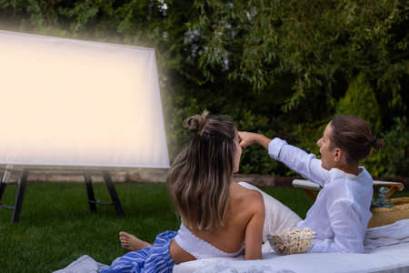 Young heterosexual couple sititng on the grass floor pointing the projector screen while in the outdoor cinemaの写真素材