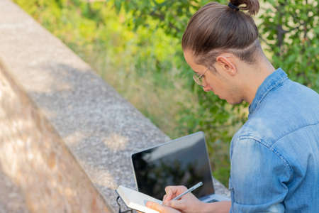 Back view of young man on a bun taking notes to create a new business online using his laptop sitting in a parkの写真素材