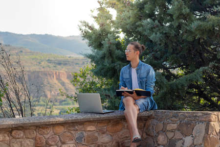 Side view of young man hard working on his online project with laptop and notebook looking to the beautiful landscapeの写真素材