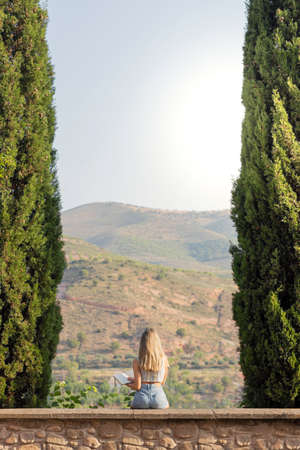 Vertical view of young woman from the back between trees holding a notebook on hands looking to the natural landscapeの写真素材