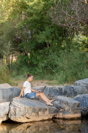 Vertical vision of a remote worker sitting on the bank of a river and working with his laptop in natureの写真素材