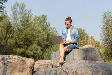 Freelancer working with his laptop from nature sitting on a stone among the trees of the forest in summerの写真素材