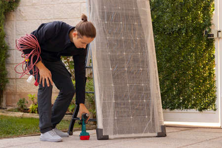 Young man worker crouched down picking up electric screwdriver to install the new solar panelの写真素材