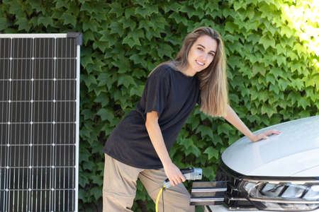 Young beautiful woman charging parked electric car from solar power station with photovoltaic panel in green backgroundの写真素材