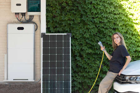 Young beautiful woman looking to camera and smiling on white electric car holding a plug and a solar system on the sideの写真素材