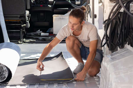 Young hipster man crouched measuring and cutting the roll isolation for the self build camper van...