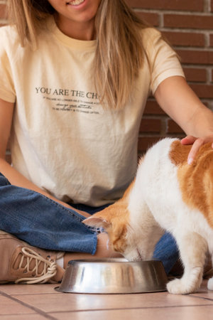 Vertical view of unrecognizable young woman smiling and sitting on the floor petting her domestic cat while eatingの写真素材