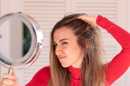 Young woman concern about her hair loss in autumn time while looking to round hand mirror in red outfitの写真素材