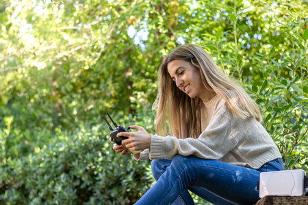 Low angle view of young blond woman smiling and happy while flying a drone with a remote controller in hands while in natureの写真素材