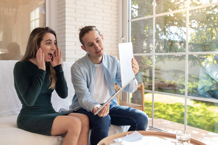 Woman surprised at something viewed on the laptop with amazed face and hands on it while man showing the screen in the living roomの写真素材