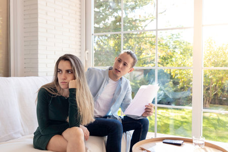 Angry young couple yelling at each other with paperwork in hands sitting in the sofa at homeの写真素材