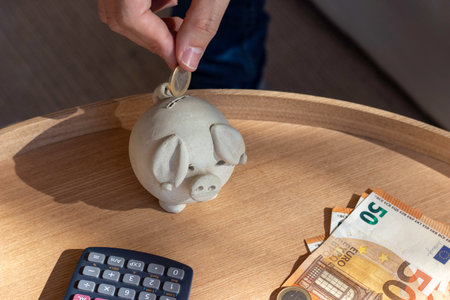 Close up top view of unrecognizable person putting euro coin into piggy bank with money bill and calculatos on the wooden table in a sunny dayの写真素材