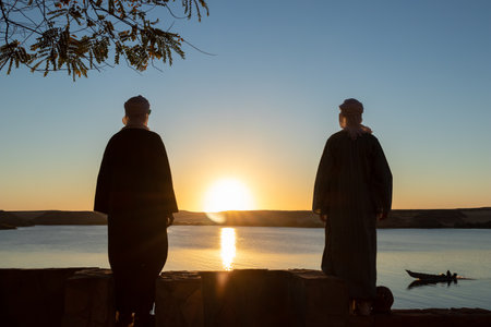 Two arab young man backward wearing djellaba and turban looking to the sunset in front of a lakeの写真素材