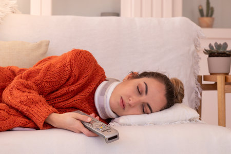 Calm young female wearing neck collar healing after trauma while holding television remote control sleeping in living roomの写真素材