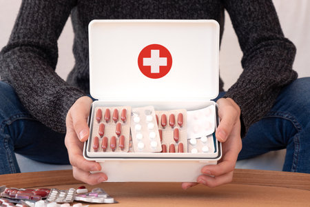 Crop male in casual clothes sitting at home and showing many medications in white first aid kit with red crossの写真素材