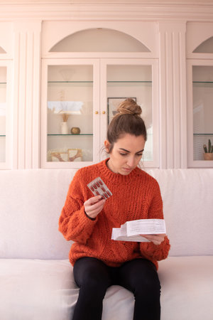 Young female in warm sweater sitting on sofa holding blister of pills while reading instructions attentivelyの写真素材