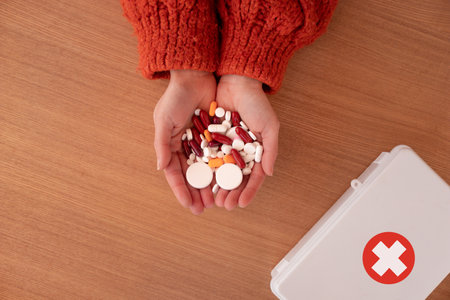 Crop female demonstrating handful of various pills and capsules on wooden table near white box with crossの写真素材