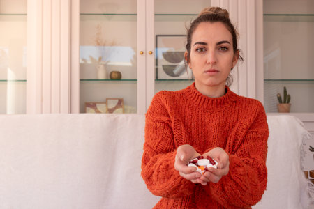 Woman in orange sweater sitting on sofa at home and showing handful of many pills while looking at cameraの写真素材