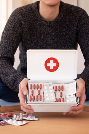 Crop male wearing gray sweater sitting at coffee table and demonstrating white box with many medicationsの写真素材